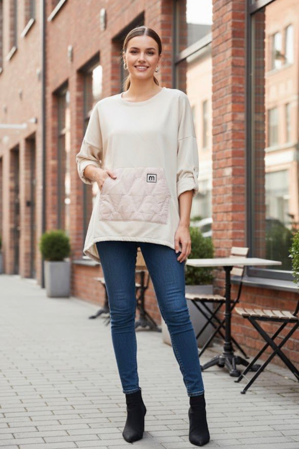 Woman wearing a beige sweater with a brand logo, blue jeans, and black boots standing on a sidewalk.